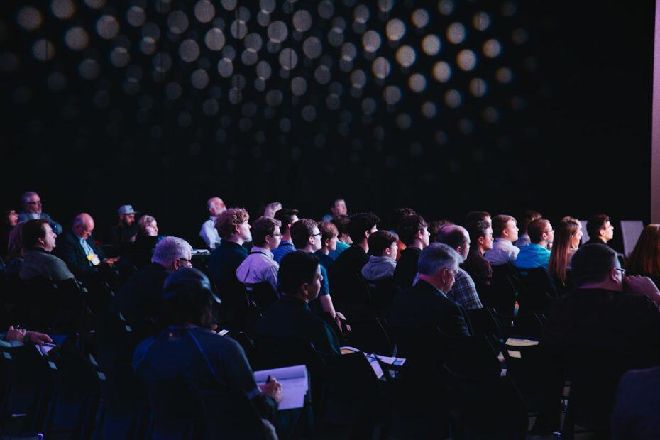 Audience attending a conference in a dark auditorium, focusing on a speaker or presentation.